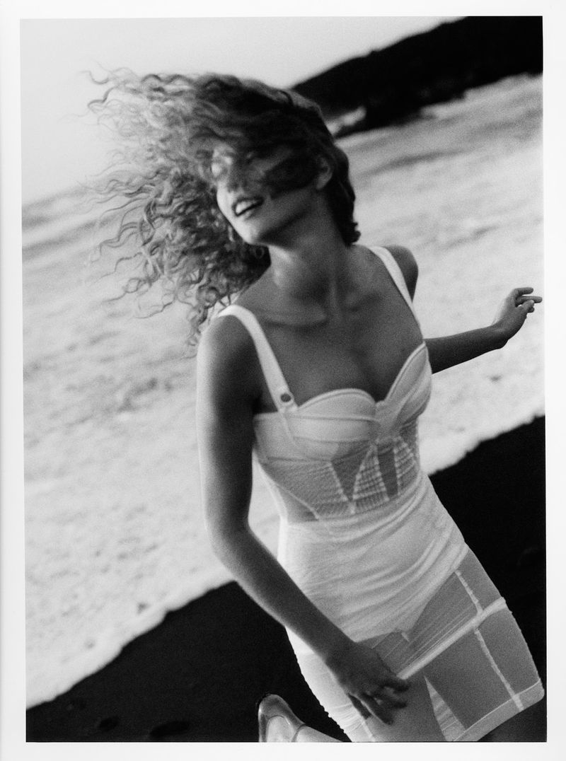 Woman in a white dress runs on a black sand beach next to crashing waves with hills in the background, a fashion editorial image.