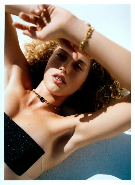 Young woman with curly hair poses with gold jewelry, including a necklace and bracelet, in a beauty or fashion editorial photo.