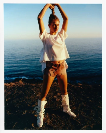 Adult female fashion model posing on a rocky beach, wearing a white blouse, tan shorts, and white boots, with the ocean in the background.