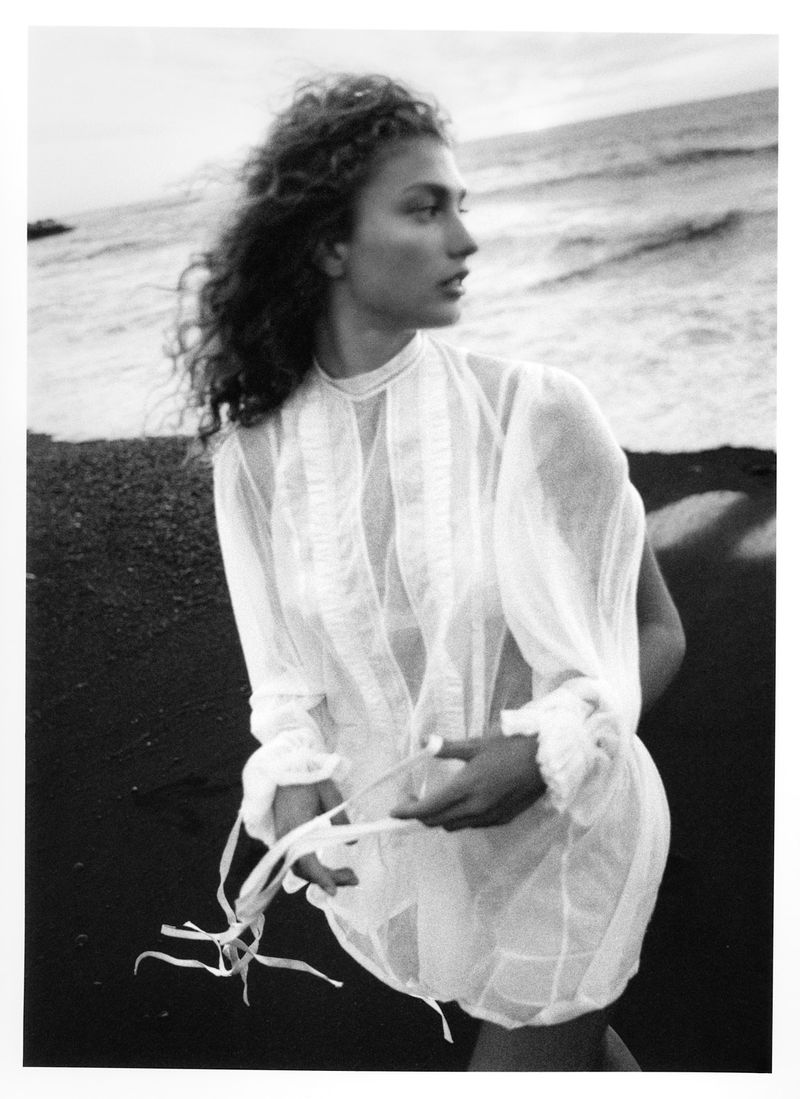 Woman with curly hair poses on a black sand beach wearing a white dress and holding a string, with the ocean in the background.