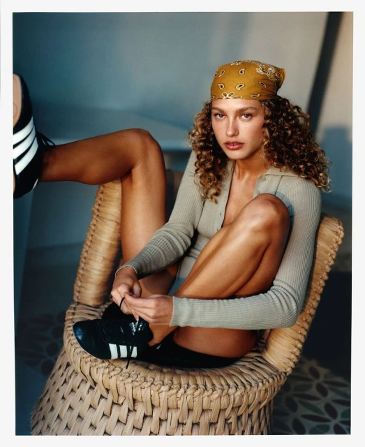 A woman with curly hair sits on a woven chair tying her Adidas shoe, wearing a tan long-sleeve and patterned headscarf in a studio.