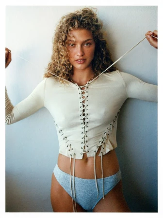 A woman with curly hair poses in a long-sleeved, lace-up top and underwear against a neutral wall, creating a fashion or beauty image.