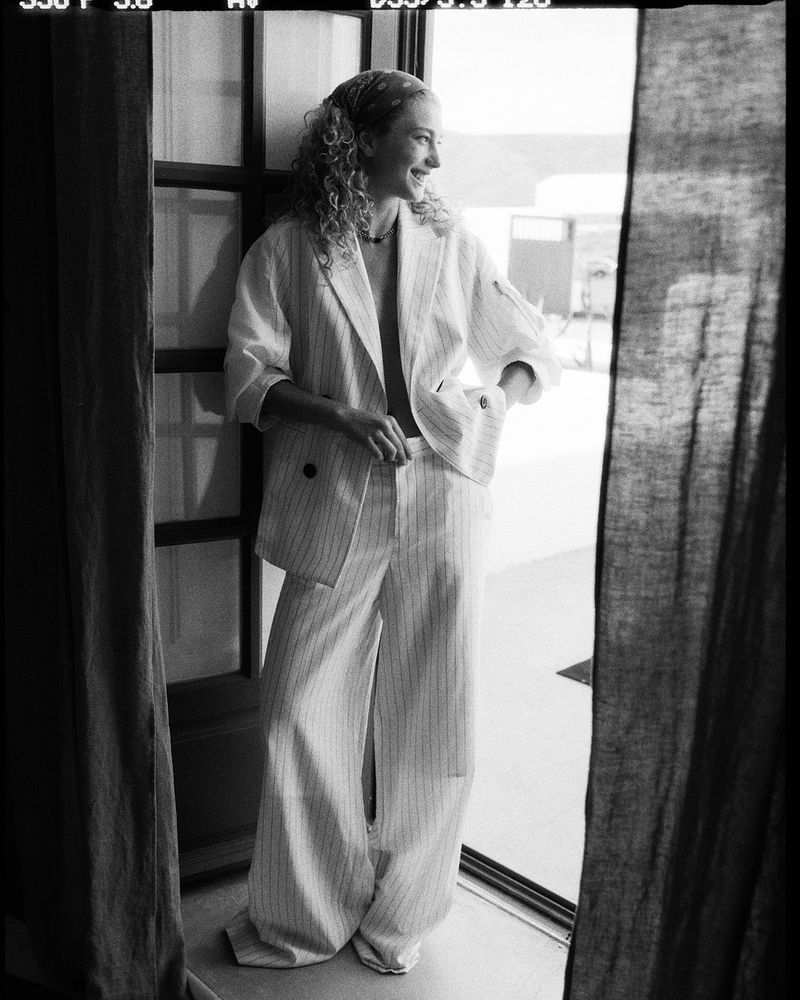 Black and white portrait of a woman in a striped suit standing in a doorway with linen curtains and curly hair, looking away.