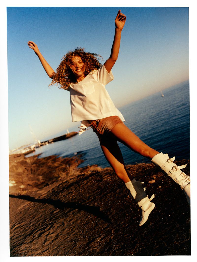 A young woman with curly hair wearing a white shirt and buckled boots jumps in the air against a backdrop of the ocean and clear blue sky.
