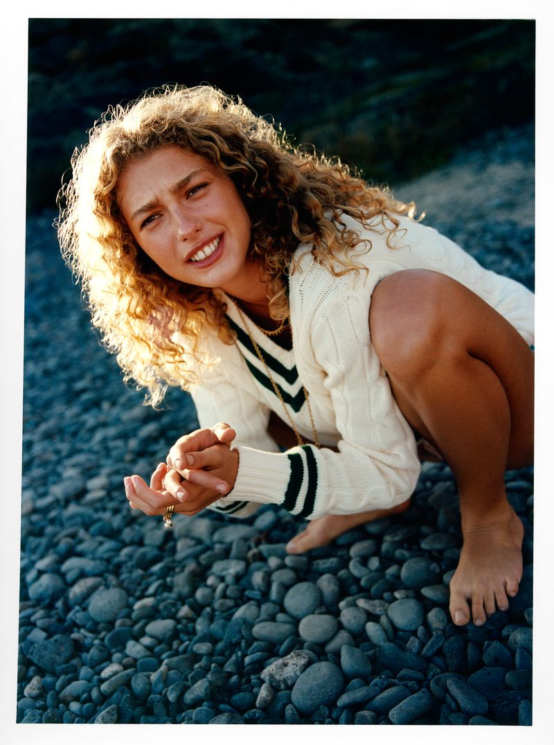 Woman with curly blonde hair in a white sweater crouches on a rocky beach, smiling in a medium shot with natural lighting.
