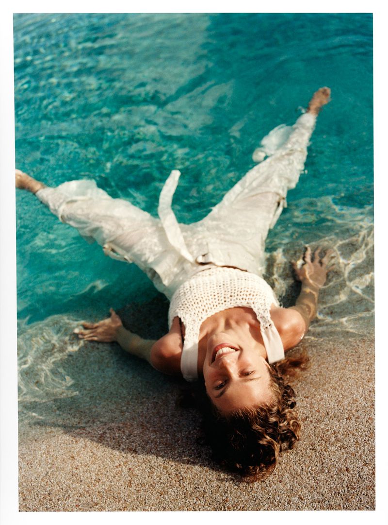 Woman in white clothing lying in shallow turquoise water on a sandy beach with a bright smile and relaxed pose, enjoying summer day.