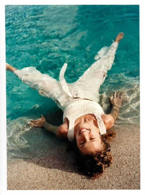 Woman in white clothing lying in shallow turquoise water on a sandy beach with a bright smile and relaxed pose, enjoying summer day.