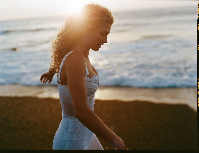 Woman in white dress walking on a sandy beach with waves in the background at sunset, casting a warm glow.