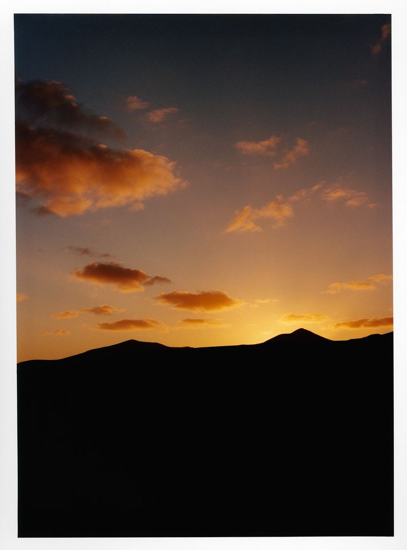 Dramatic sunset landscape featuring silhouetted mountain range against a sky with colorful orange and peach-colored clouds.
