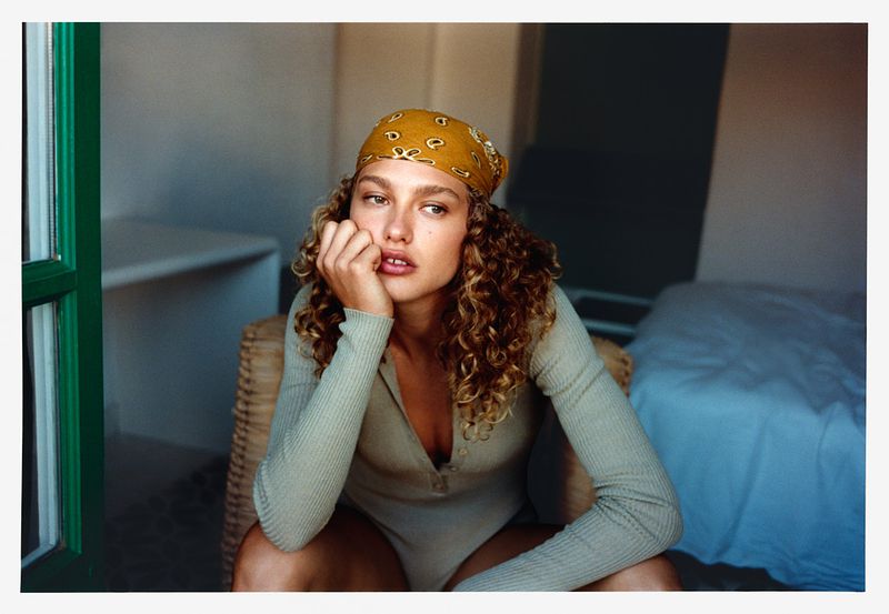 Indoor portrait of an adult female with curly brown hair wearing a yellow patterned bandana, posing in a beige bodysuit on a wicker chair.