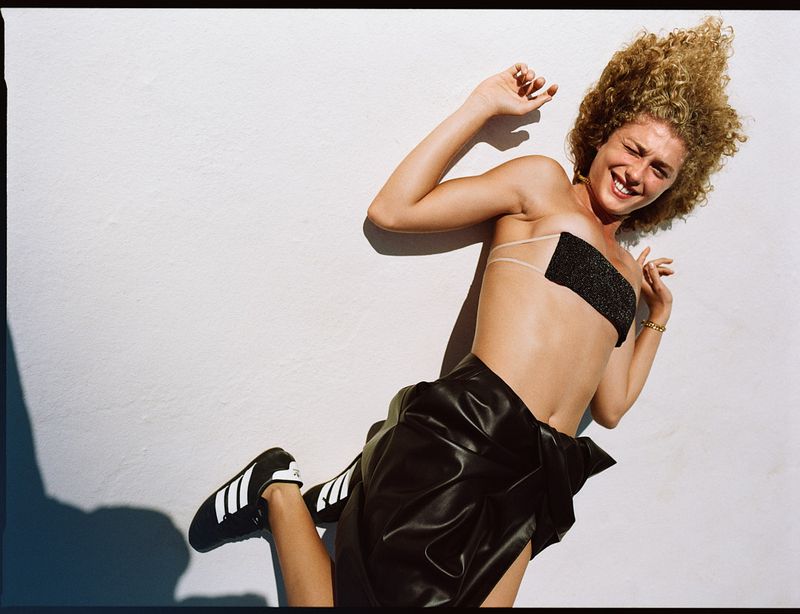 Young woman with curly hair poses in shiny black bandeau top and skirt with Adidas sneakers against white wall backdrop.