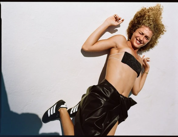 Young woman with curly hair poses in shiny black bandeau top and skirt with Adidas sneakers against white wall backdrop.