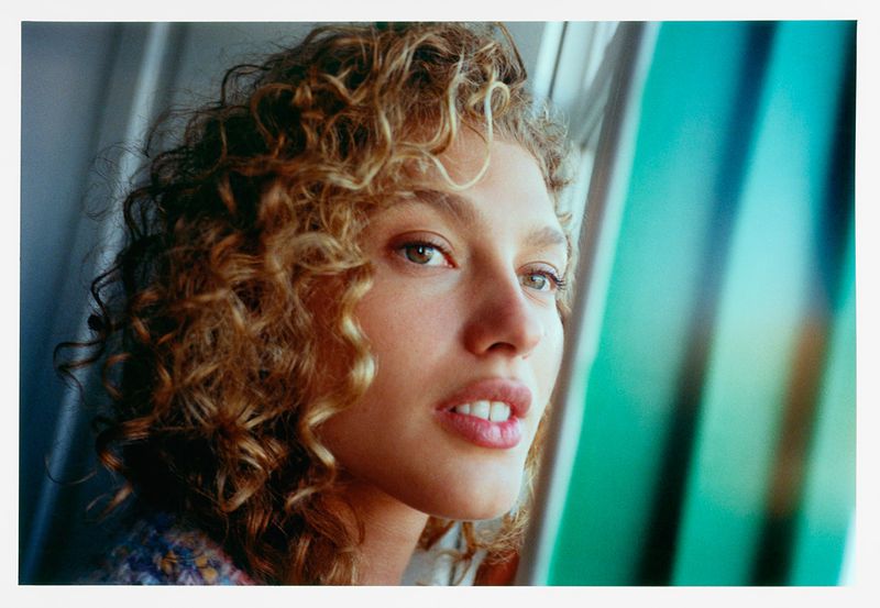 Close-up portrait of a curly-haired adult female looking slightly upward, with indoor natural lighting and neutral expression.