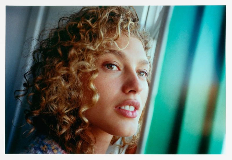 Close-up portrait of a curly-haired adult female looking slightly upward, with indoor natural lighting and neutral expression.
