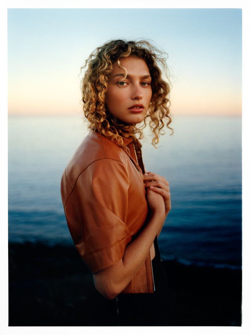 Woman with curly hair poses in front of ocean wearing leather jacket in a medium shot with soft, diffused sunset lighting.