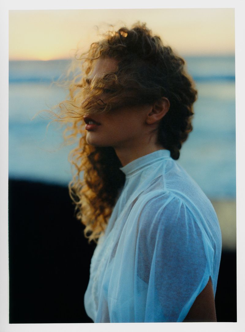 A young woman with curly red hair, wearing a white top, stands on a beach with the sun setting over the ocean in the background.