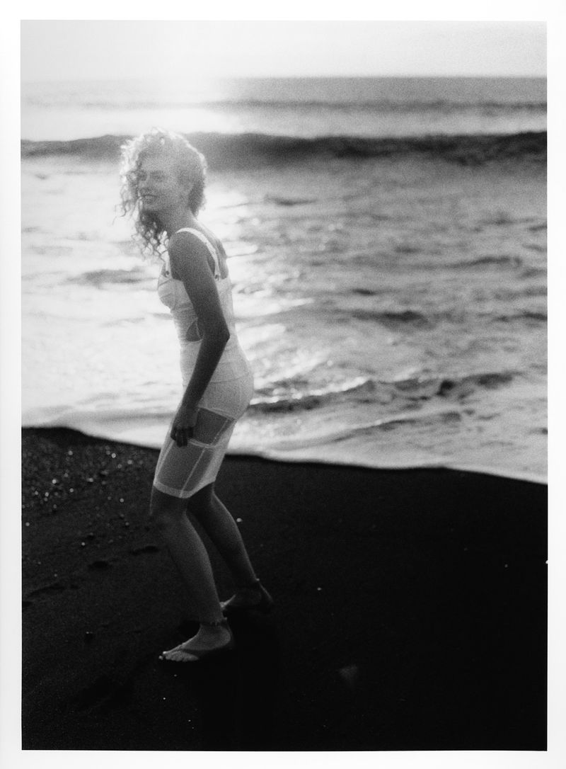 Monochrome portrait of a smiling woman in a vintage swimsuit walking along a dark sand beach as waves crash gently in the background.