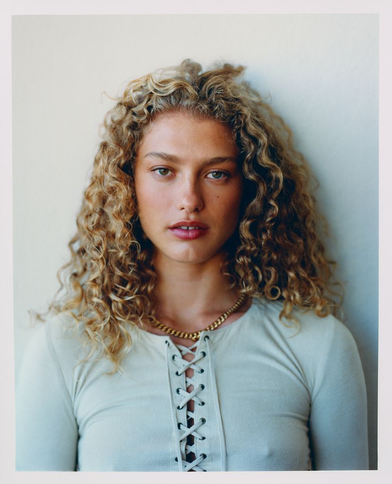 Close-up portrait of an adult woman with curly blonde hair wearing a white, laced top and gold necklace, in front of a neutral background.