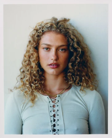Close-up portrait of an adult woman with curly blonde hair wearing a white, laced top and gold necklace, in front of a neutral background.