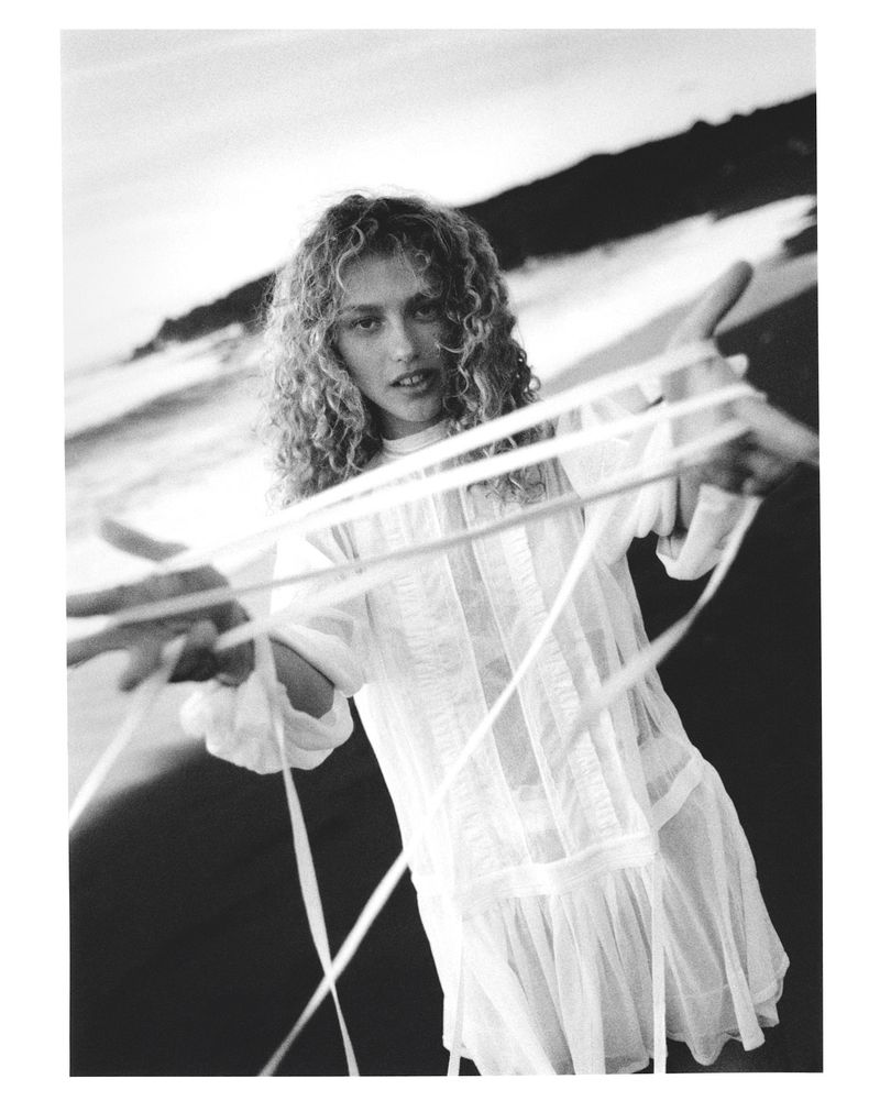 Black and white fashion shot of a young adult female with curly blonde hair in a short white dress, holding white ribbons on a beach.