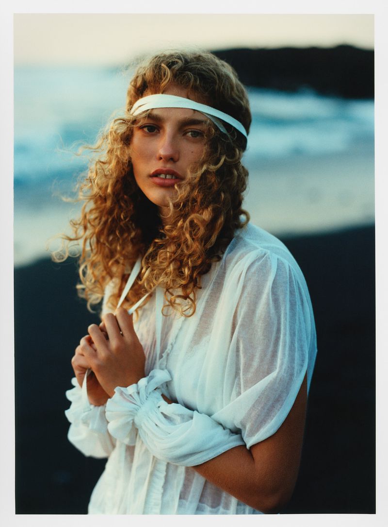 Portrait of woman with blonde curly hair wearing white dress and headband on beach with ocean backdrop during daytime.