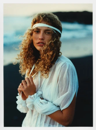 Portrait of woman with blonde curly hair wearing white dress and headband on beach with ocean backdrop during daytime.