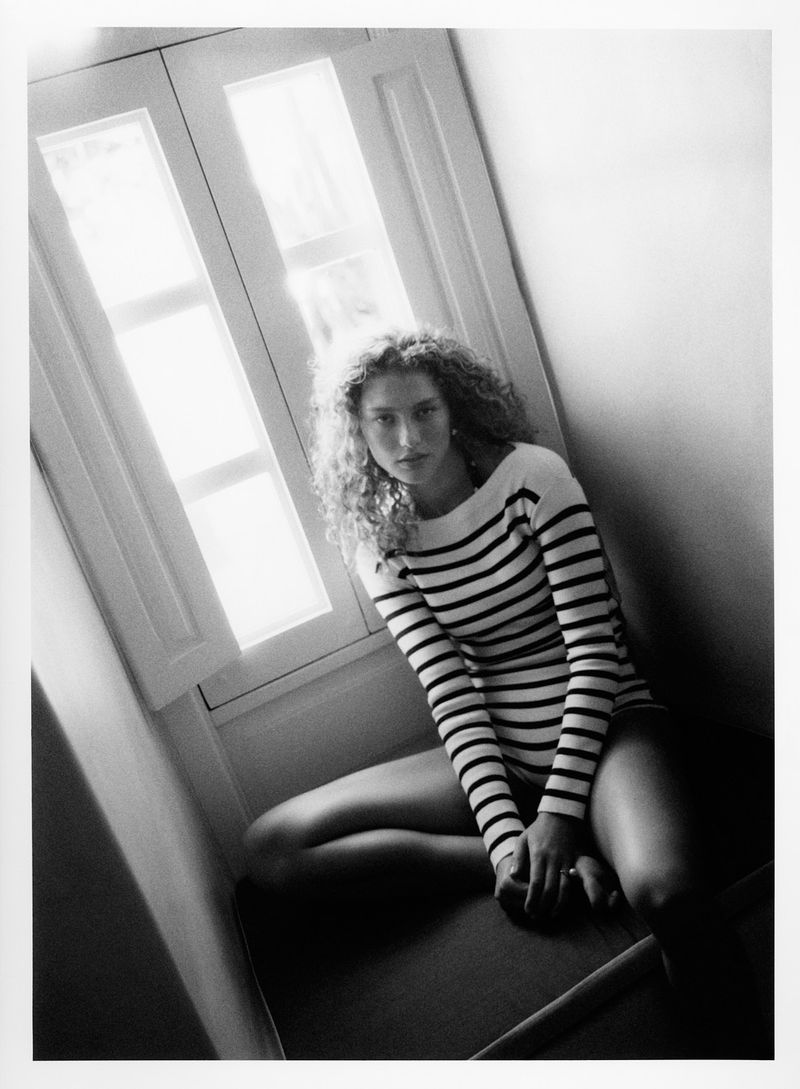 Black and white portrait of a woman with curly hair, wearing a striped shirt and shorts, sitting near a window with natural lighting.