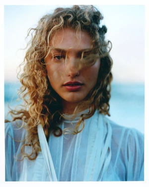 Close-up portrait of model Dorit Revelis with blonde curly hair wearing a white top outdoors against a blurred background.
