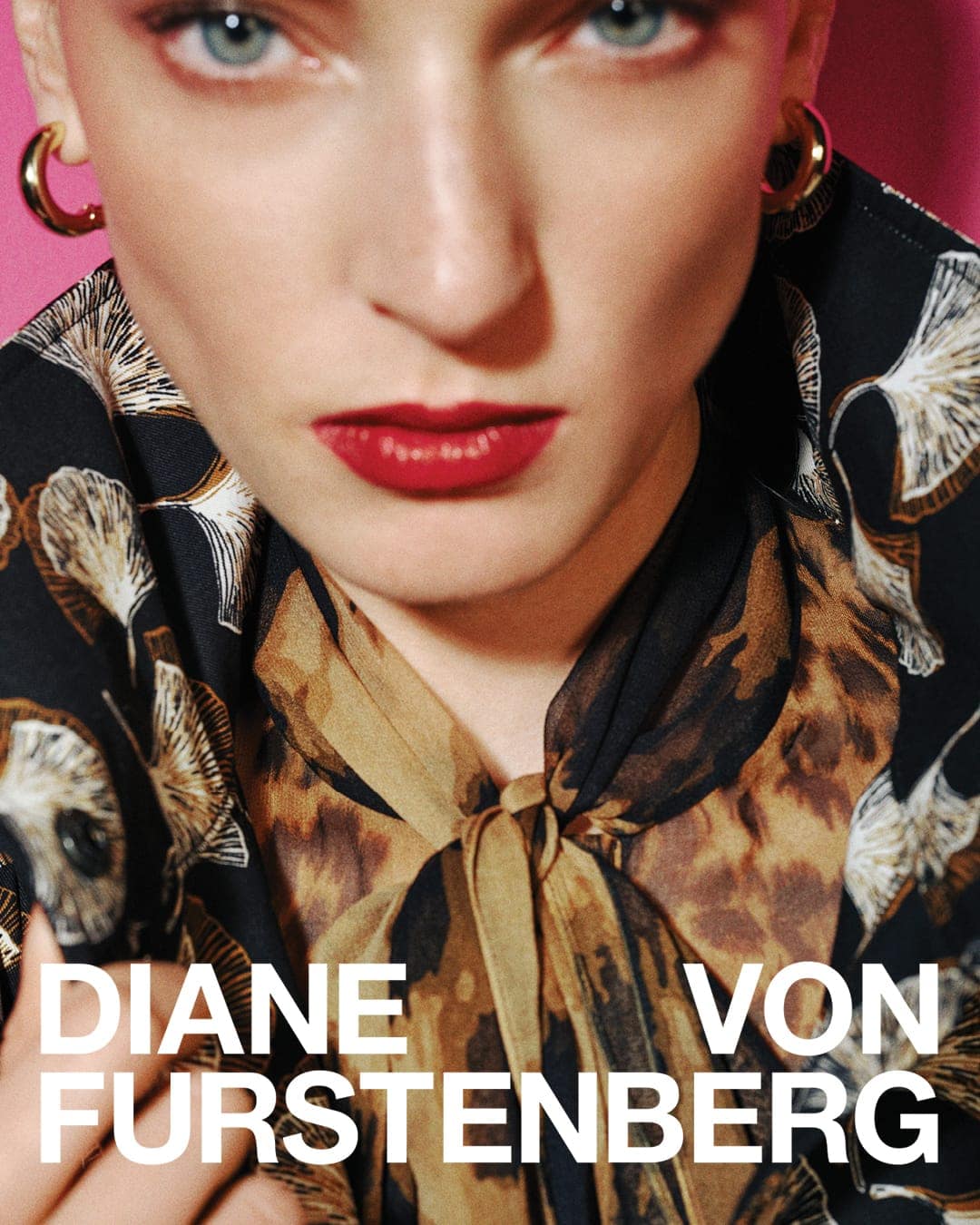 Close-up portrait of a model with red lips and gold hoop earrings wearing a patterned blouse against a pink background