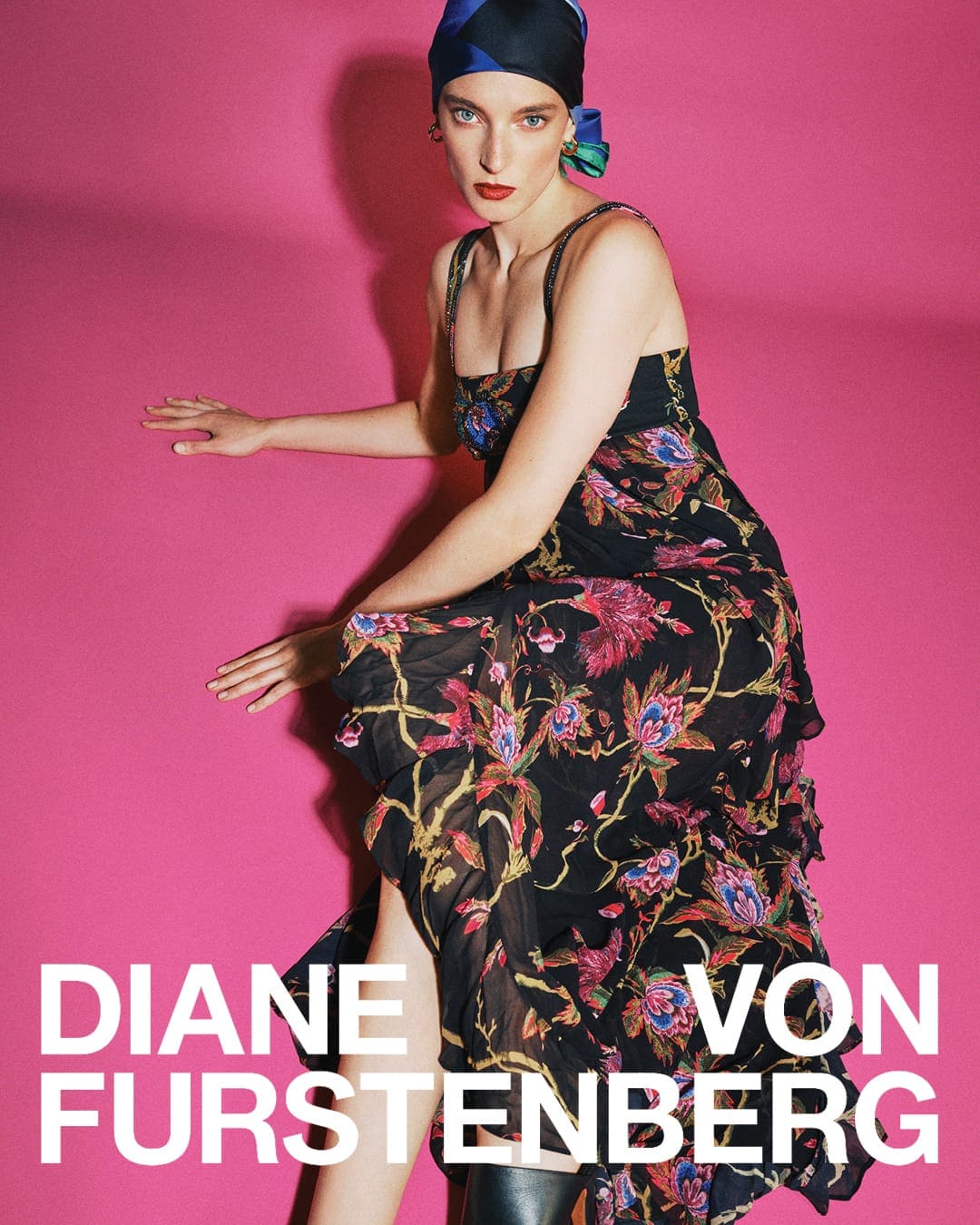Model in a floral print maxi dress and patterned headscarf seated against a bright pink background for Diane von Furstenberg.