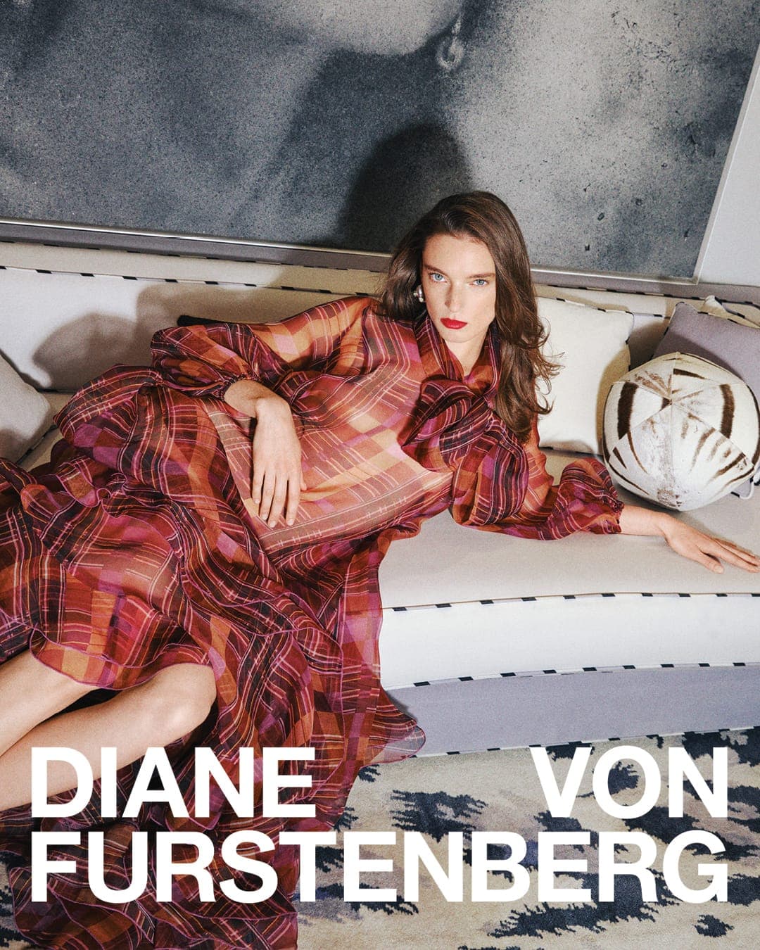 Model lying on a sofa wearing a long red plaid dress in an indoor setting for Diane von Furstenberg
