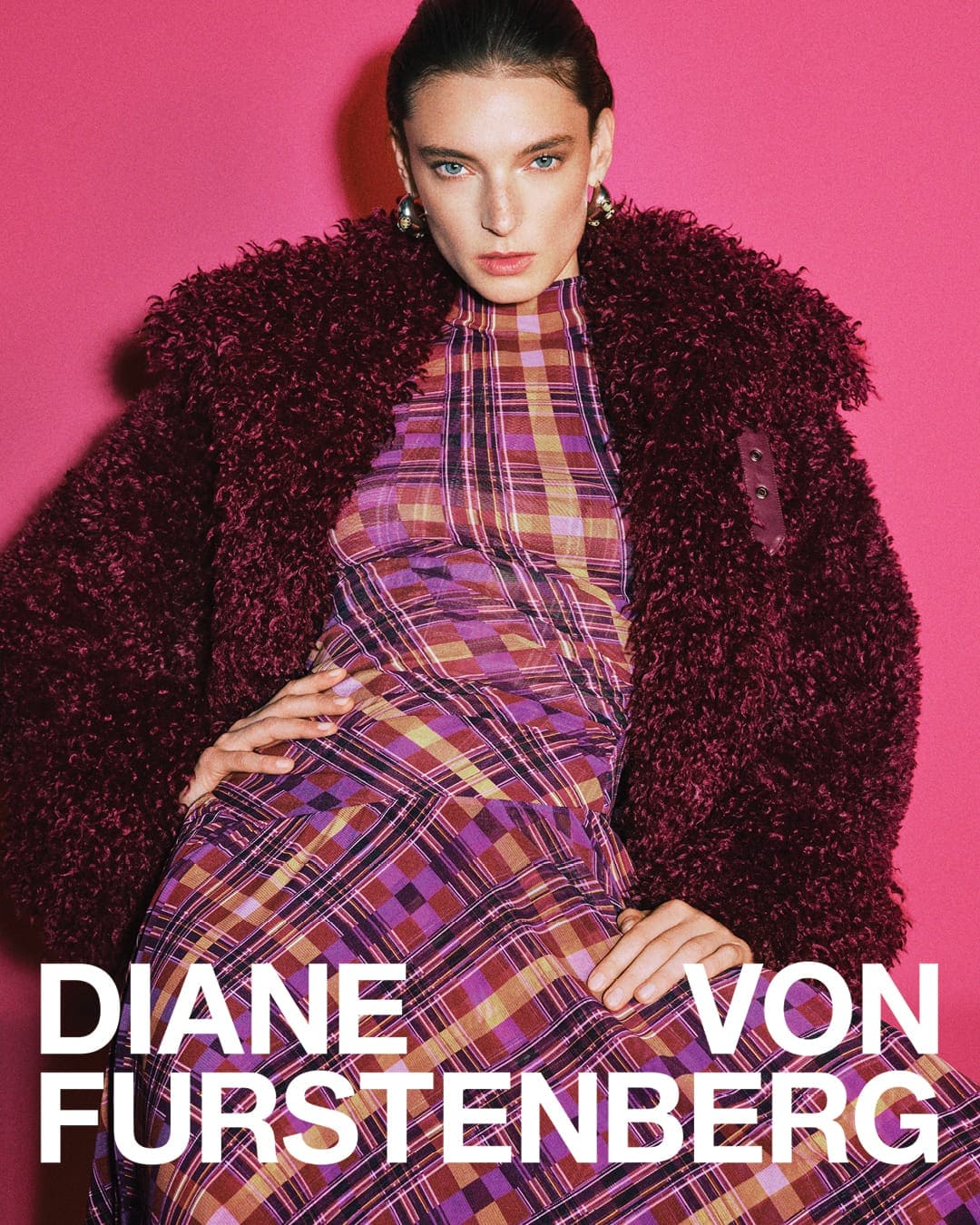 Model in burgundy textured coat and plaid dress posing against vibrant pink background for Diane von Furstenberg Fall 2025 campaign