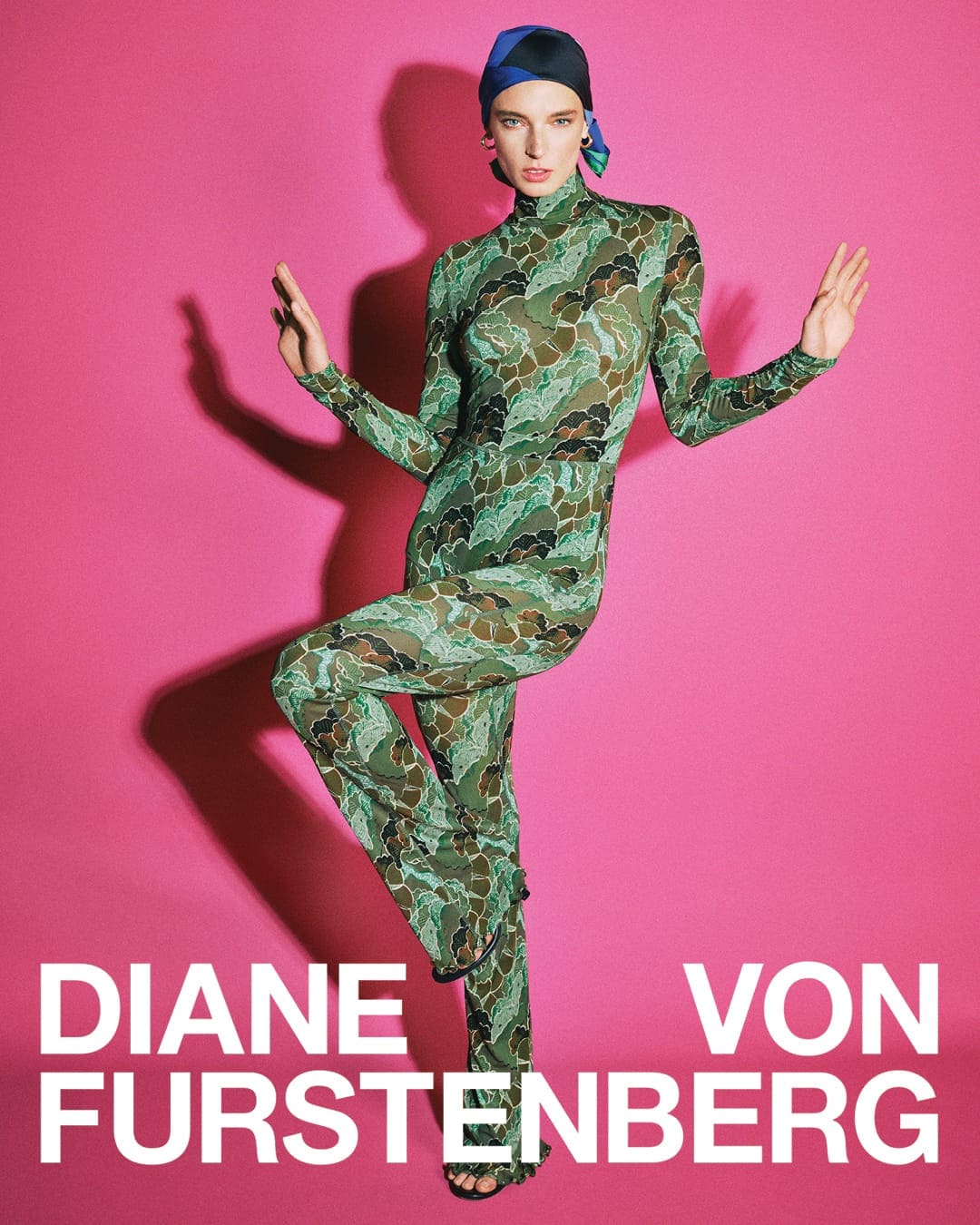 Model in green patterned jumpsuit and headscarf posing against a bold pink backdrop for Diane von Furstenberg campaign