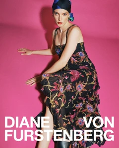 Model in a floral print maxi dress and patterned headscarf seated against a bright pink background for Diane von Furstenberg.