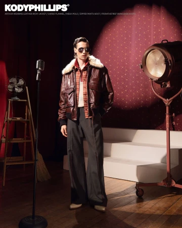 Model wearing dark sunglasses, brown leather jacket with fur collar, and dark trousers standing on a stage with a studio spotlight.