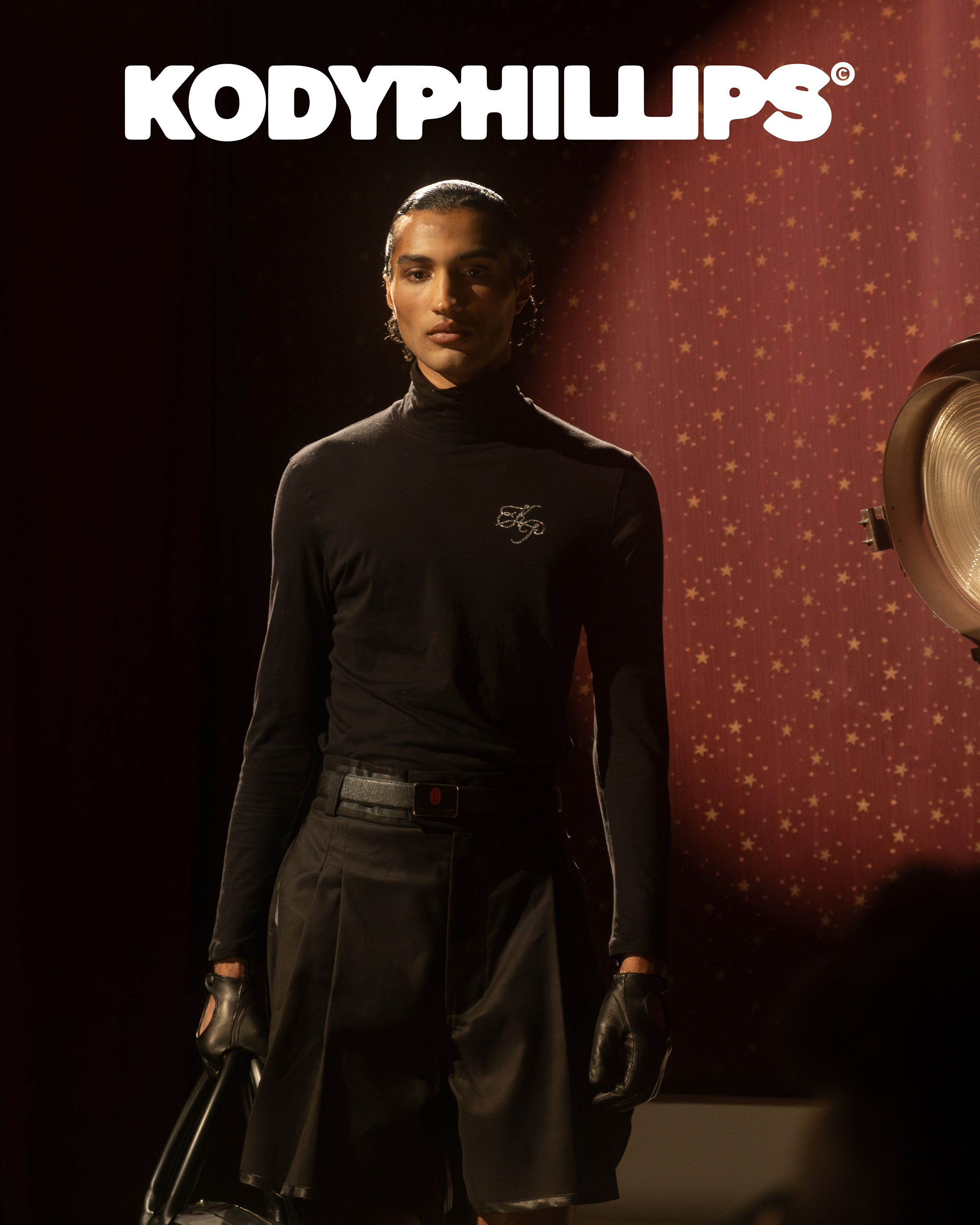 Model in black turtleneck and dark shorts wearing gloves against a patterned red backdrop with a studio spotlight.