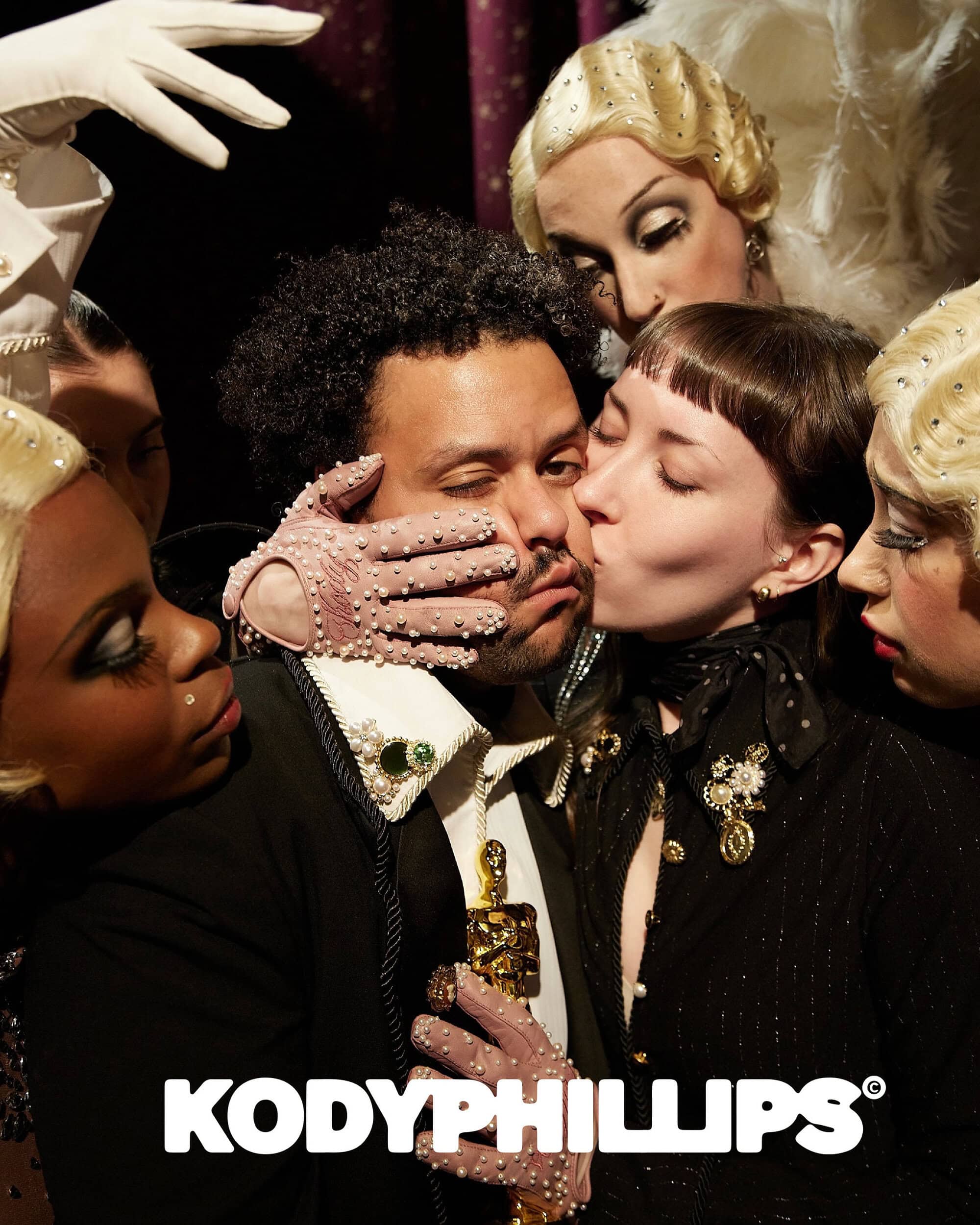 Man surrounded by multiple women in elaborate vintage hairstyles and embellished gloves in a dramatic staged portrait.
