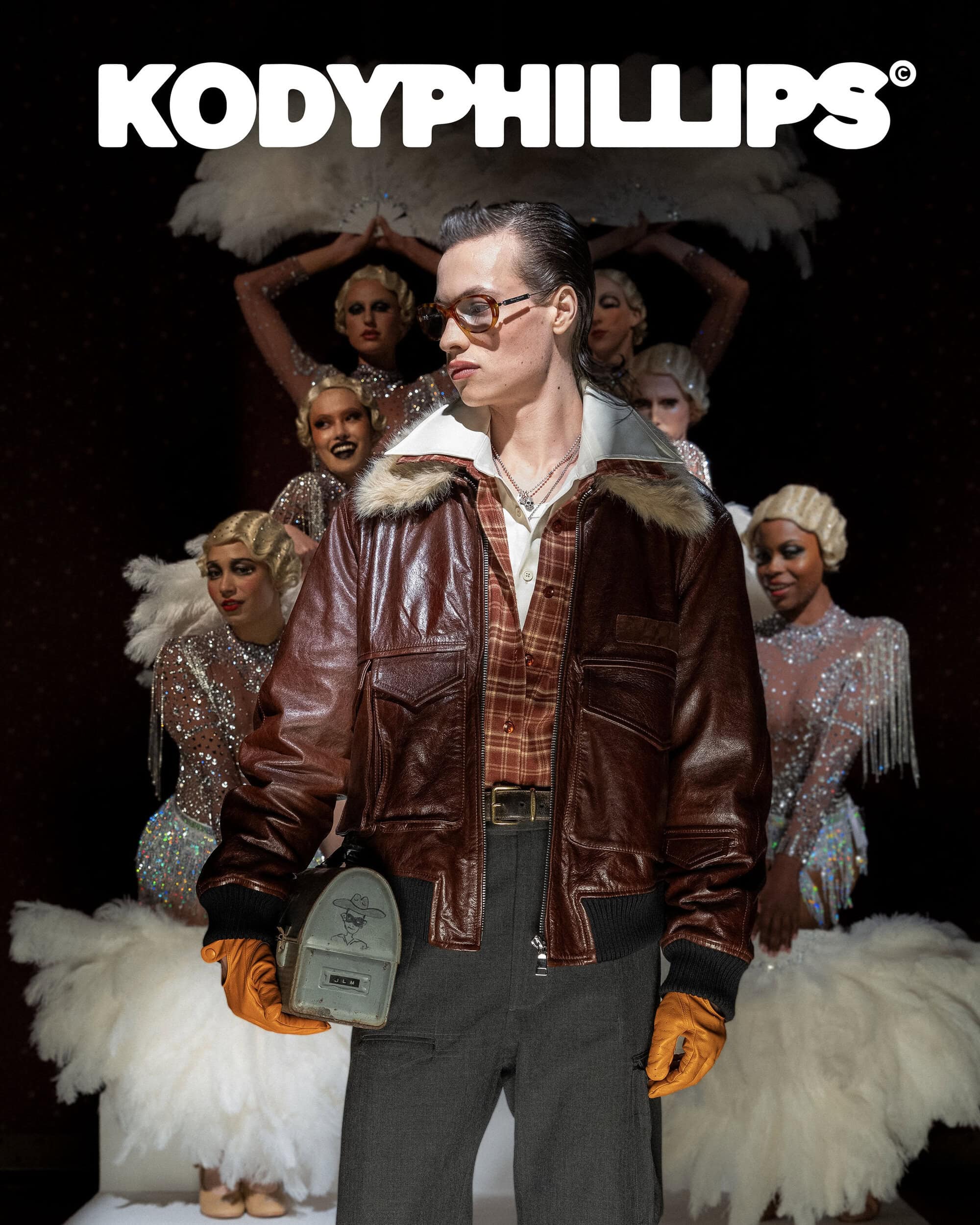 A man wearing a brown leather flight jacket and orange gloves holding a small metallic box, with a group of female performers behind.