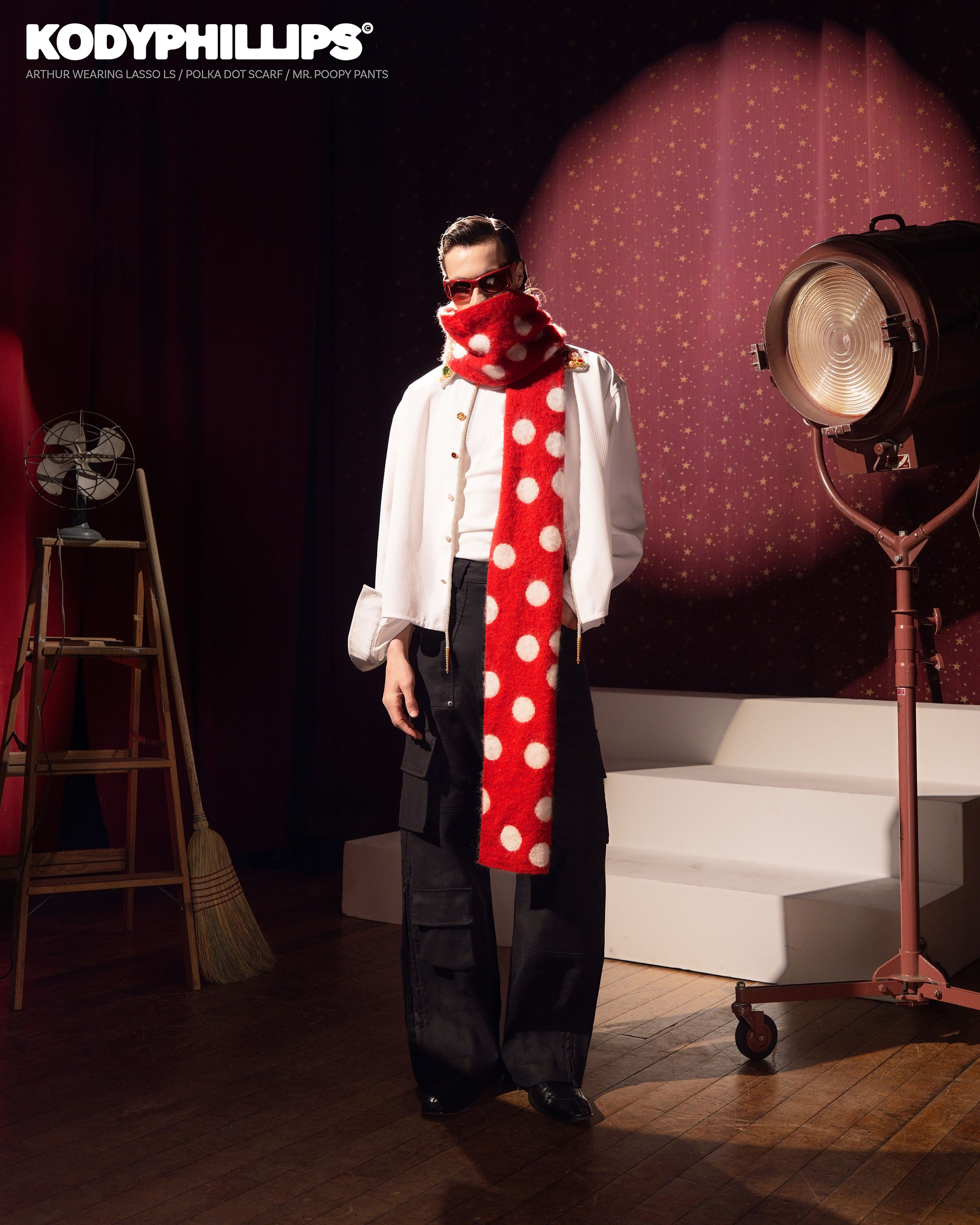 Model wearing a red and white polka dot scarf, white shirt, and dark trousers standing on a wooden floor in a studio set.