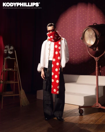 Model wearing a red and white polka dot scarf, white shirt, and dark trousers standing on a wooden floor in a studio set.