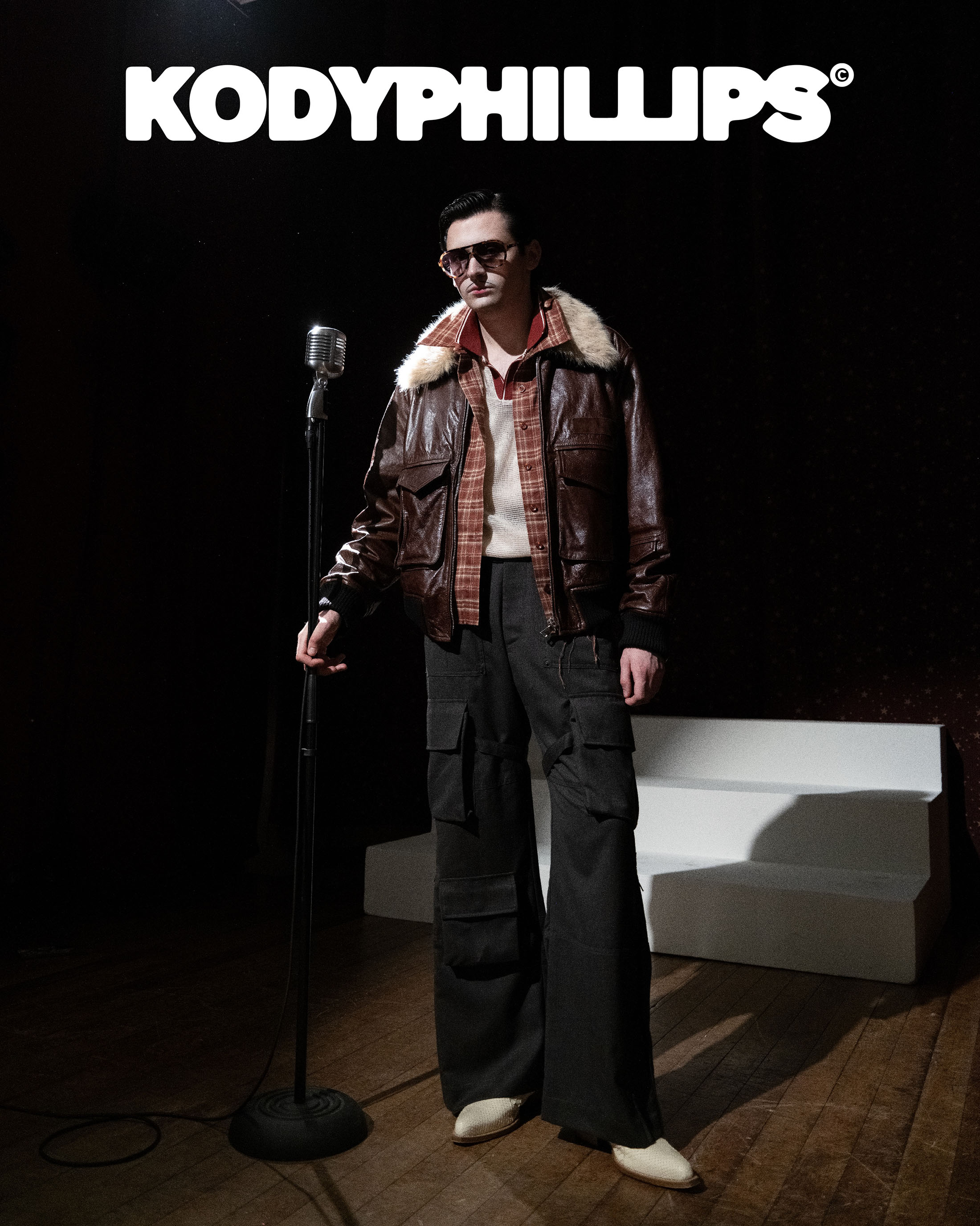 Man wearing a brown leather jacket and sunglasses standing next to a vintage microphone in a dark studio setting