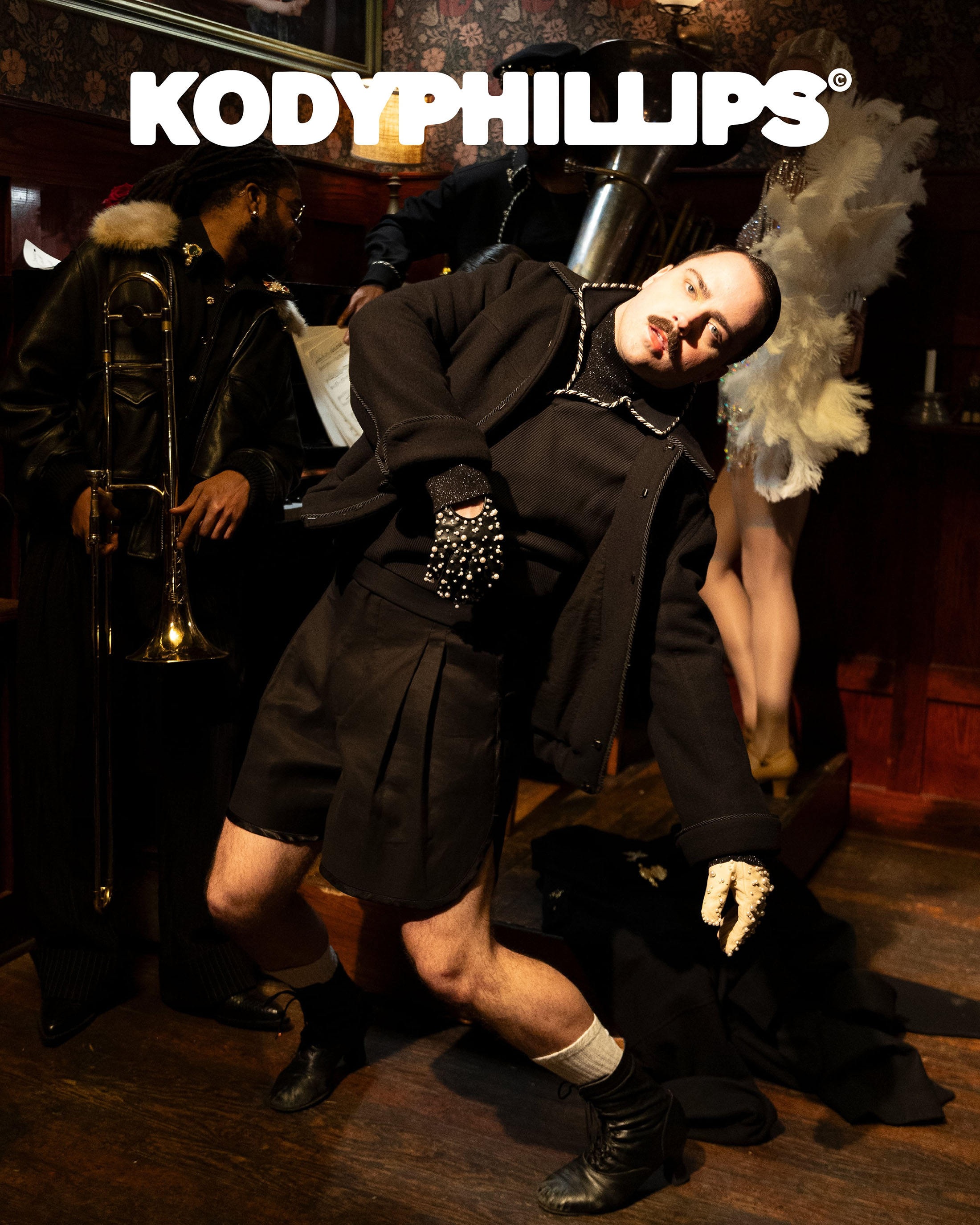 Model leaning back dramatically wearing a black jacket and shorts in a dimly lit bar setting with musicians.