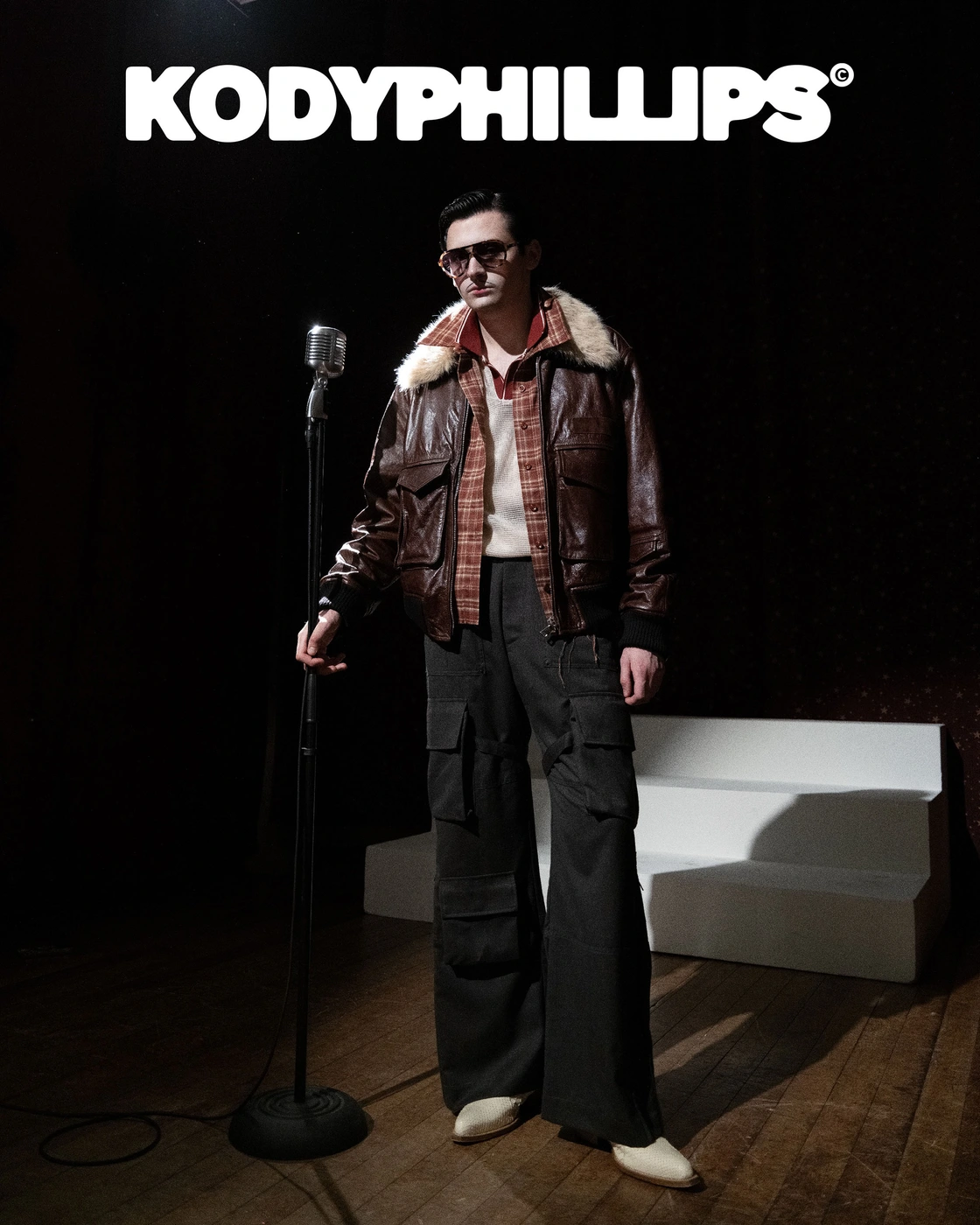 Man wearing a brown leather jacket and sunglasses standing next to a vintage microphone in a dark studio setting