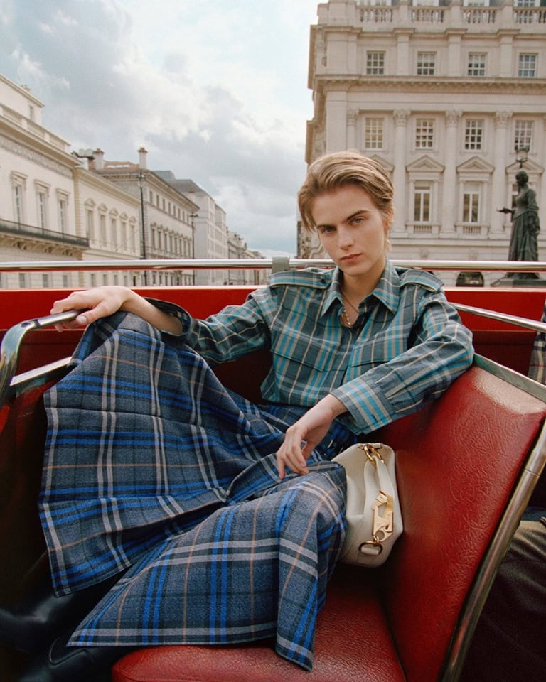 Model seated on the upper deck of a red double-decker bus wearing blue plaid clothing against a city street background.