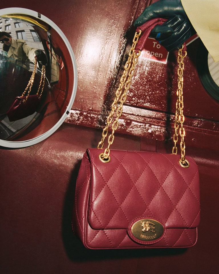 Burgundy quilted leather handbag with gold chain strap held by a gloved hand against a dark red interior.