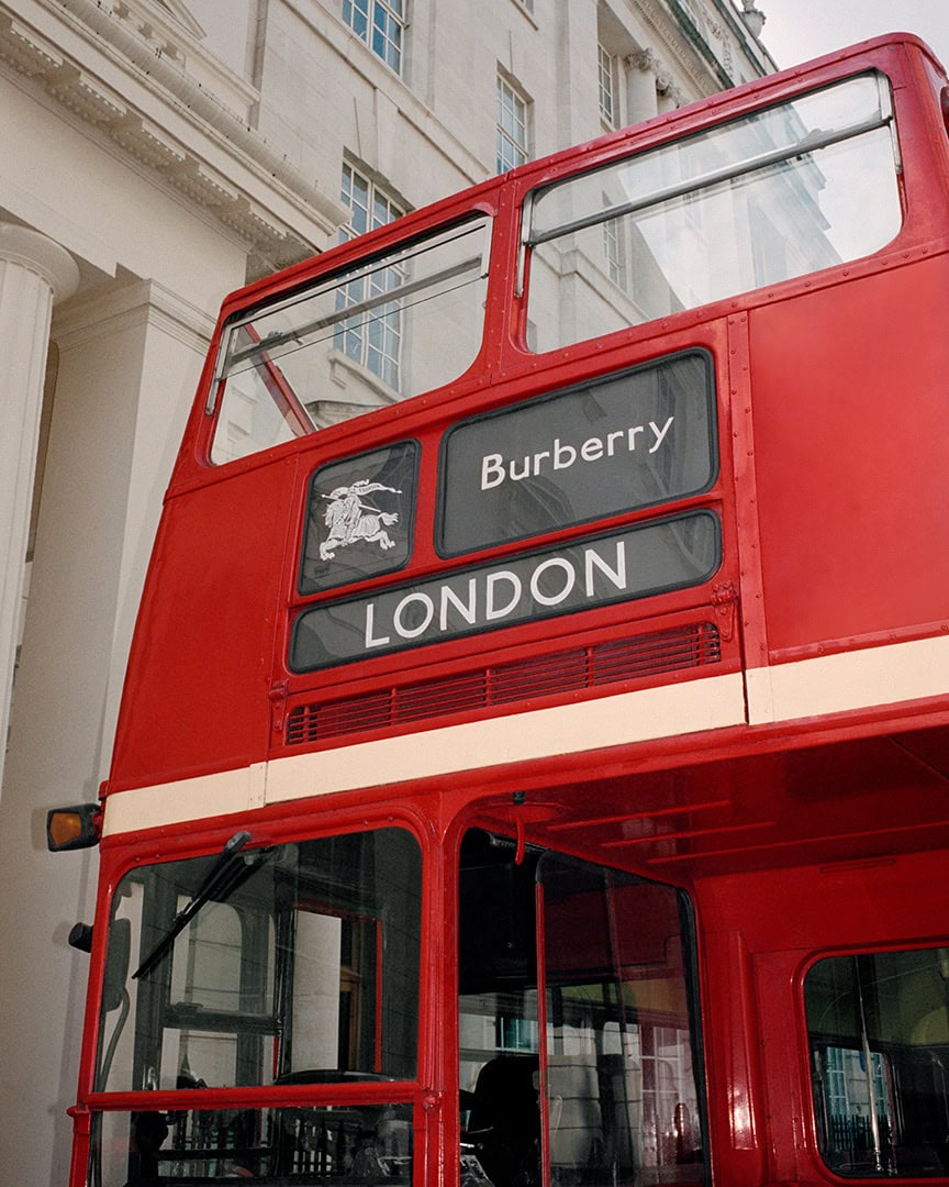 Close-up of a red double-decker bus with Burberry branding parked in front of a classical stone building exterior.