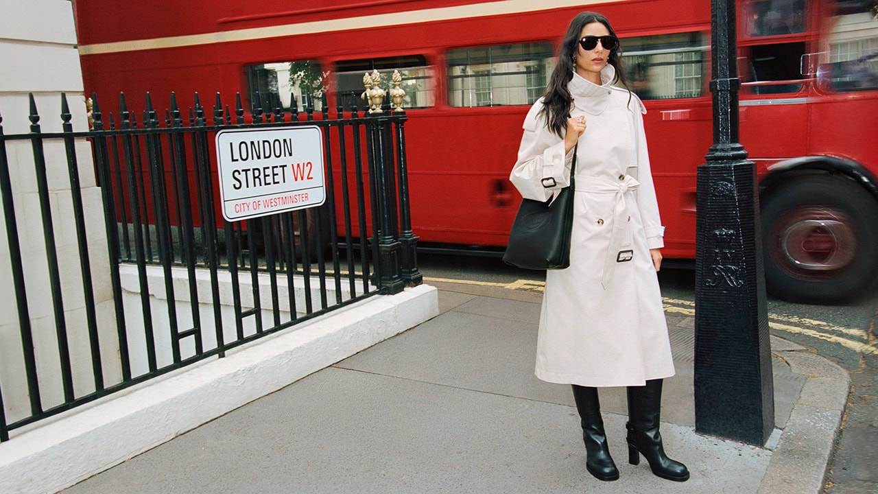 Model in cream trench coat and black boots standing on a London street in front of a red double-decker bus.