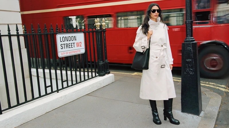 Model in cream trench coat and black boots standing on a London street in front of a red double-decker bus.