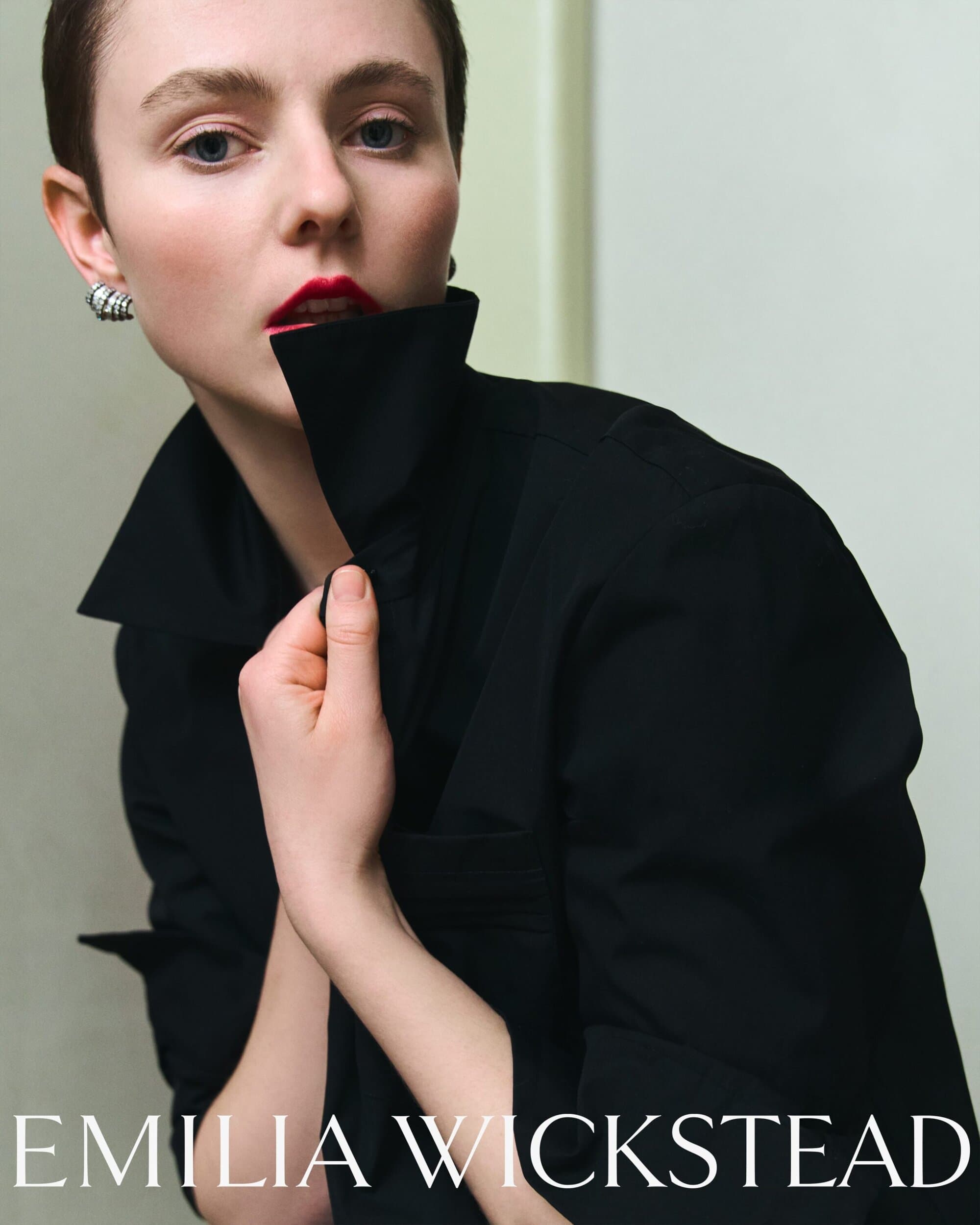 Thomasin McKenzie in a black collared shirt holding up the collar with red lipstick against an interior background