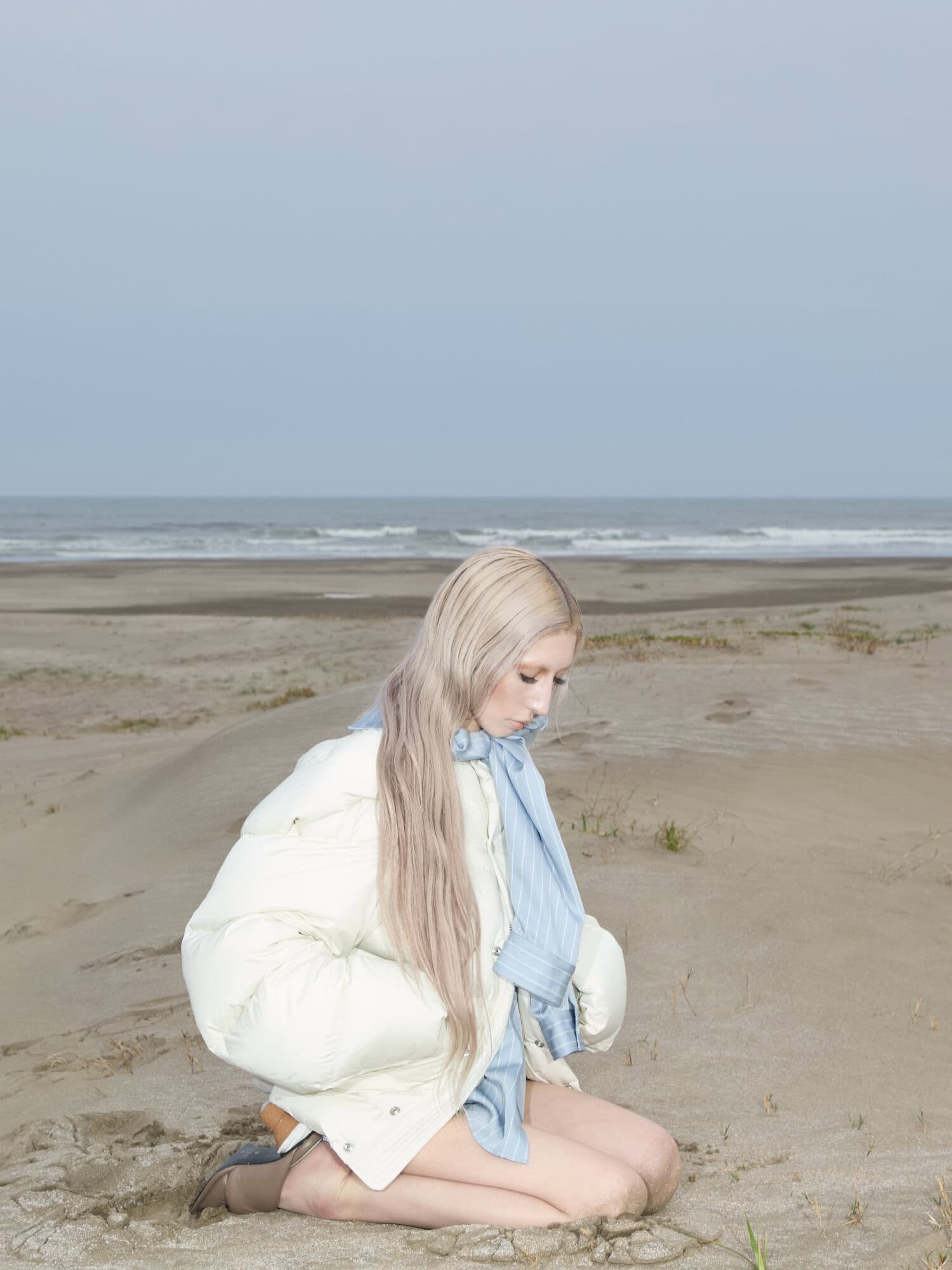 A blonde model kneeling on a sandy beach wearing a white puffer jacket and a light blue scarf.
